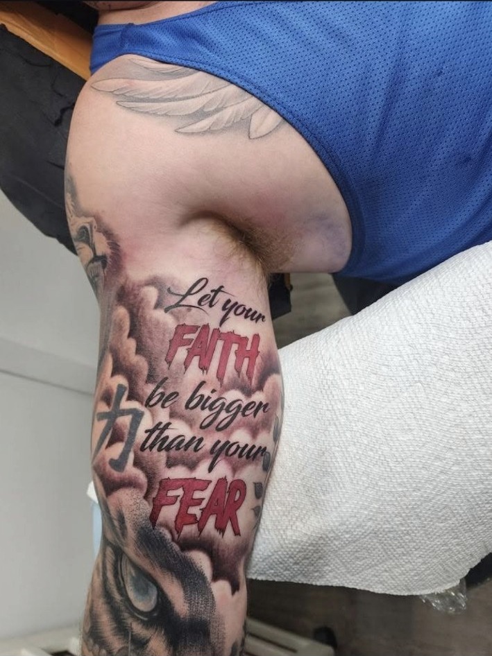 Black and grey faith over fear upper arm tattoo with red lettering by Miguel